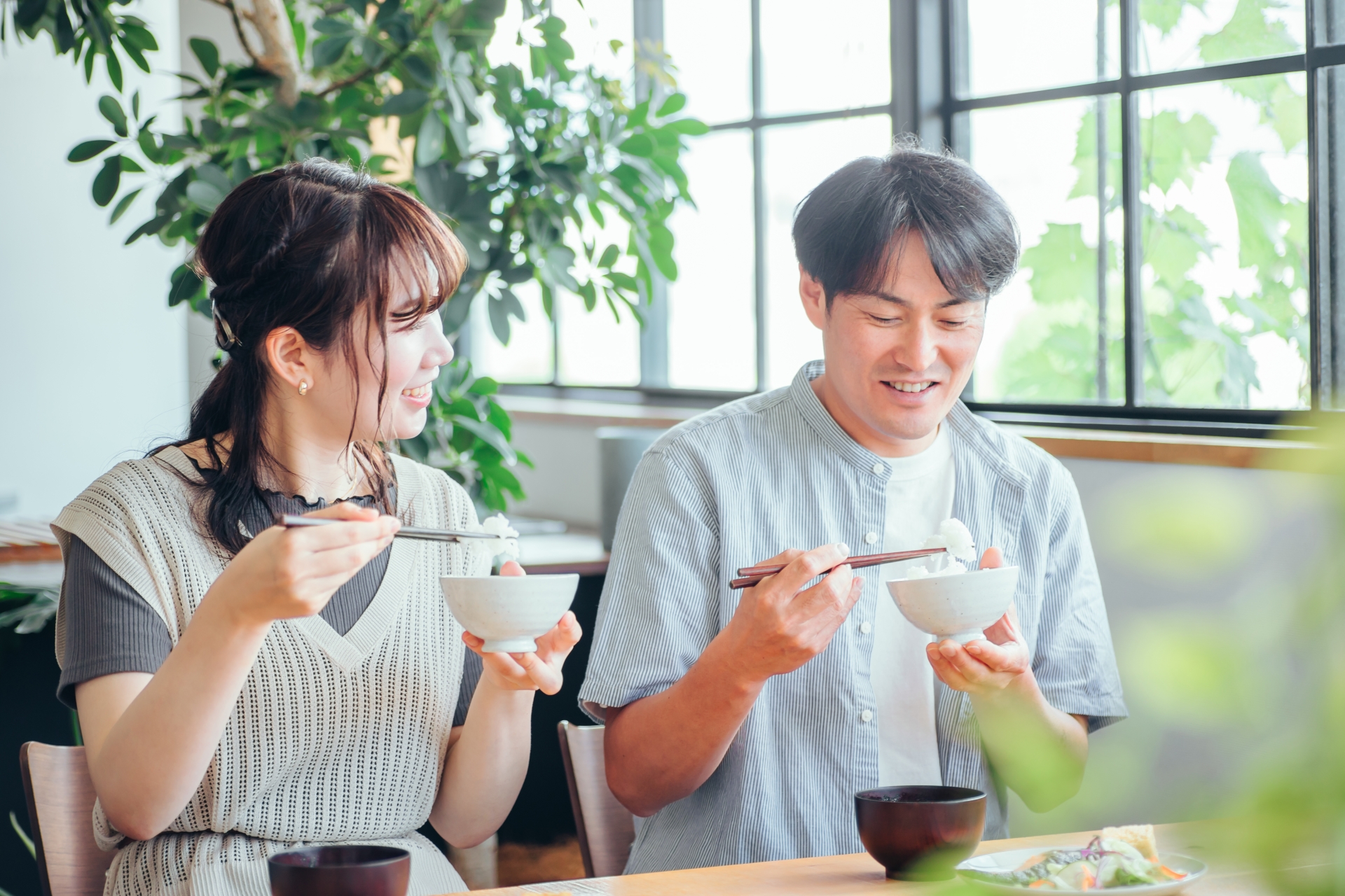 Two people holding rice bowls in one hand and chopsticks in the other at a table.