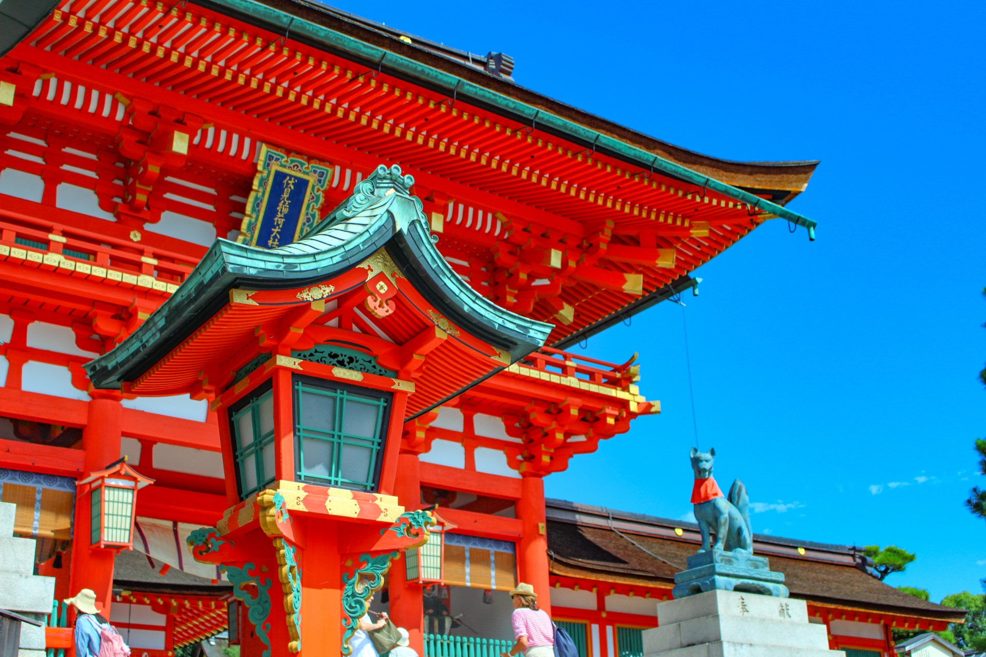 Vermilion shrine architecture, a decorative lantern and a fox statue at Fushimi Inari beneath a blue sky.