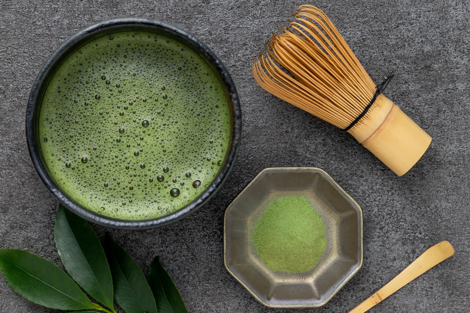 A bowl of whisked matcha beside matcha powder and a bamboo chasen.