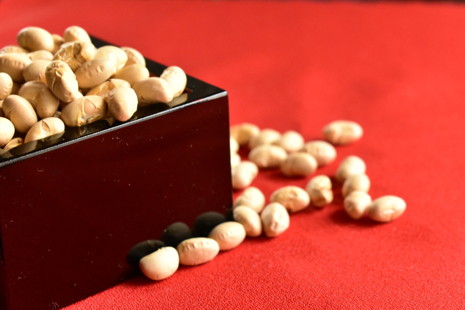 Roasted soybeans fill a dark wooden masu, with loose beans scattered across a red surface.