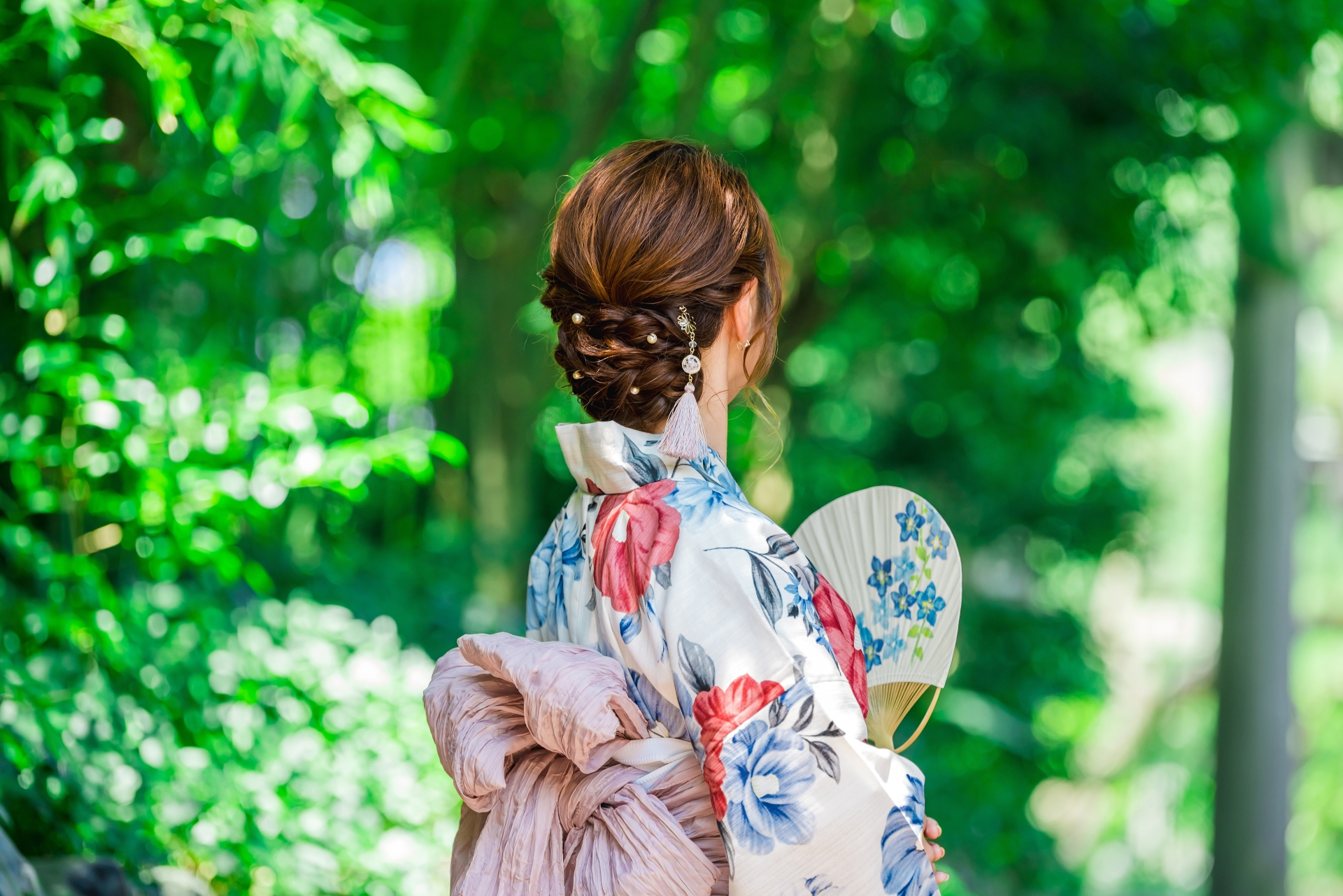 A woman in a floral yukata seen from behind among green trees, holding a folding fan.