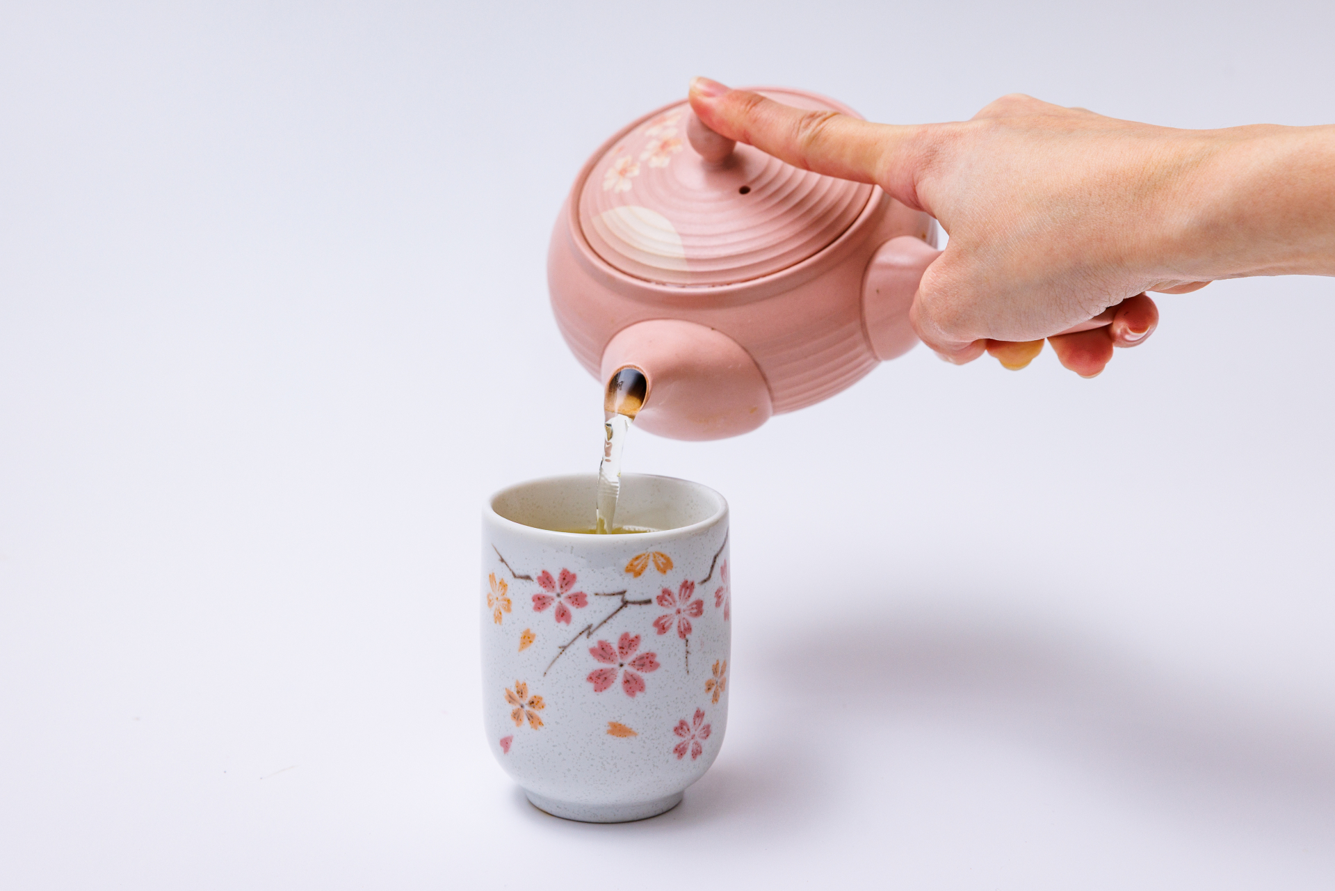 A hand pouring tea from a pink side-handled kyusu into a teacup.