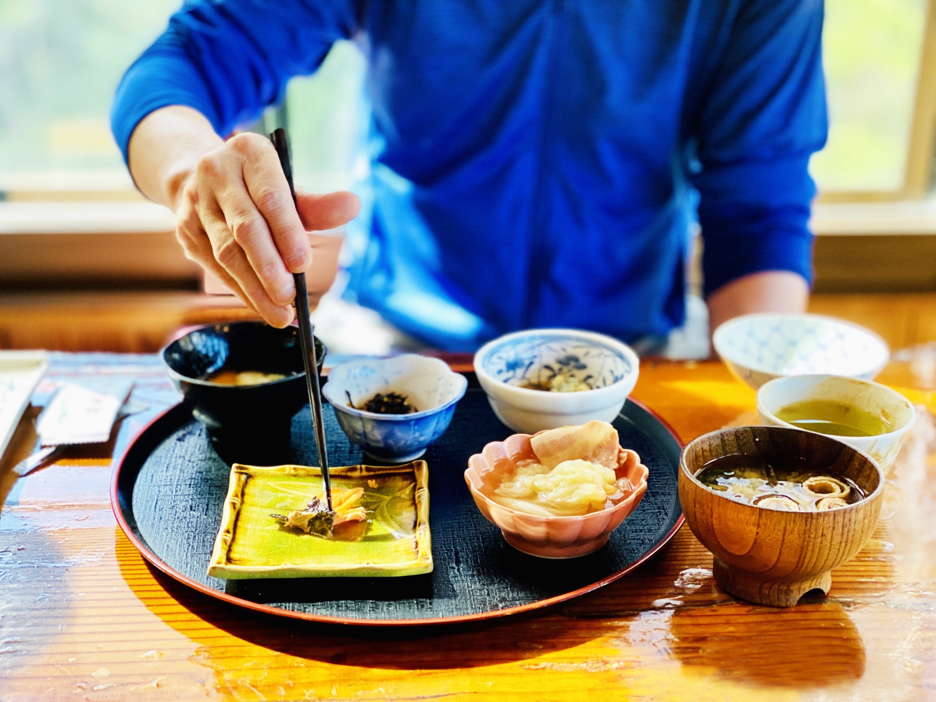 A person in a blue shirt eating a Japanese meal served in varied bowls and dishes.